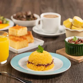 Café da manhã com sabores regionais Mesa de café da manhã com um prato da sobremesa regional cartola, uma tigela de pães de queijo e uma fatia de bolo de rolo, com frutas e café ao fundo.