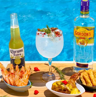 Petiscos e drinks na piscina do Salinas Maceió Bandeja na beira da piscina com uma variedade de petiscos, como camarão e isca de peixe, e bebidas, incluindo um drink de gin e uma cerveja Corona, com a água azul da piscina ao fundo.