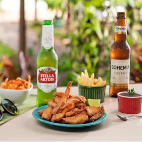Petiscos e cervejas no Salinas Maceió Mesa com um prato de isca de peixe frita, batata frita e molhos, ao lado de duas garrafas de cerveja, Stella Artois e Bohemia. A cena se passa em uma área externa com plantas ao fundo.