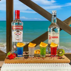 Drinks e shots no Salinas Maceió Bandeja de madeira com uma garrafa de vodka Smirnoff e uma de cachaça Ypióca ao lado de quatro shots coloridos enfeitados com laranja, morango e kiwi, tendo o mar de Maceió como cenário de fundo.
