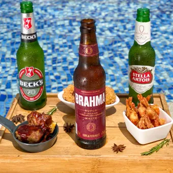 Cervejas e petiscos na piscina do Salinas Maceió Bandeja de madeira na beira da piscina com três cervejas, Beck's, Brahma Duplo Malte e Stella Artois, e porções de petiscos, incluindo camarão frito e costelinha barbecue, com a água azul da piscina ao fundo.