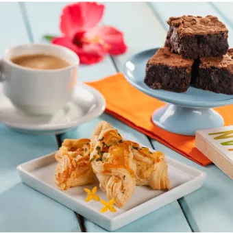 Café da tarde no Salinas Maceió Mesa azul com uma xícara de café, um prato de folhados salgados e um suporte com brownies de chocolate. Uma mão segura a xícara e uma flor de hibisco decora o ambiente, representando uma pausa para o café no resort.