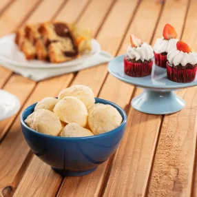 Lanche da tarde no Salinas Maceió Mesa posta em uma superfície de madeira, com uma tigela azul de pães de queijo em destaque, além de cupcakes com morango, fatias de bolo mesclado e uma xícara de café preto ao fundo.