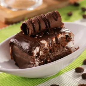 Sobremesas e café no Salinas Maceió Fatia de bolo de chocolate com recheio cremoso e uma bebida de café com chantilly ao fundo. A cena, que representa uma sobremesa no resort, é decorada com grãos de café espalhados sobre a mesa.
