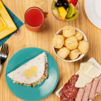 Café da tarde completo no Salinas Maceió Vista de cima de uma mesa de café da tarde completa no resort, com tapioca, pão de queijo, frios, fatias de bolo, salada de frutas, suco e uma xícara de café.
