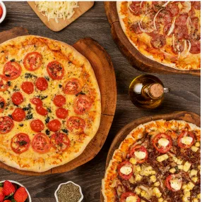 Noite da Pizza no Salinas Maceió Vista de cima de uma mesa de madeira com várias pizzas, incluindo margherita, calabresa e uma pizza doce de chocolate com morango, cercadas por ingredientes frescos e um cortador de pizza.