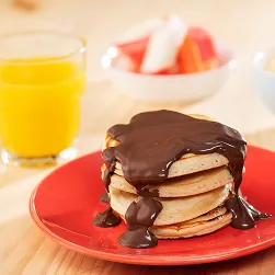 Café da manhã doce Pilha de panquecas com calda de chocolate em prato vermelho, com suco de laranja, frutas, pão de queijo e iogurte com granola ao fundo sobre mesa de madeira.