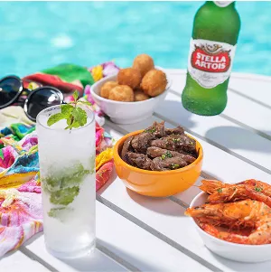 Petiscos all inclusive Mesa branca à beira da piscina com mojito no copo alto, cumbuca de carne em tiras, bolinhos fritos, camarões e uma mão segurando uma cerveja Stella Artois; óculos e canga colorida ao lado.