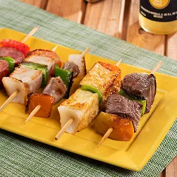 Petiscos e cerveja Prato amarelo com espetinhos variados de carne, queijo, linguiça e legumes sobre jogo americano verde, ao lado de panelinha com arroz, tigela de farofa e garrafa de cerveja Corona bem gelada sobre mesa de madeira.