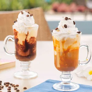 Lanches e cafés Duas taças de vidro com cafés gelados cobertos com chantilly e grãos de café, uma com calda de chocolate e outra com caramelo; ao fundo, ambiente de resort desfocado. Grãos de café espalhados e guardanapo azul sobre a mesa.
