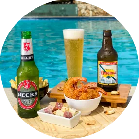 Destino incrível merece os melhores petiscos, cerveja gelada e tudo que tem direito Petiscos variados com camarões empanados, batatas fritas, queijo com linguiça e cerveja gelada sobre bandeja de madeira à beira da piscina.