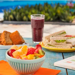Lanches saudáveis Tigela de salada de frutas, copo de suco roxo, sanduíches e fatias de bolo sobre mesa à beira da piscina.