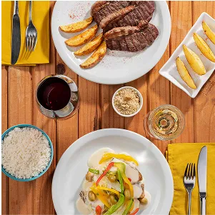 Title: Jantar all inclusive Bife grelhado com batatas e peixe ao molho com pimentões, arroz, farofa e taças de vinho sobre mesa de madeira.
