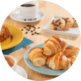 Férias em família pede croissant quentinho, cappuccino com espuma e cookies pra fechar a tarde com sabor. Uma mesa com cappuccino, croissants, bolo, cookies com pratos azul, branco e amarelo. Acompanhado de um café preto e flores decorativas.