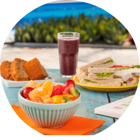 Café da tarde que combina com céu azul e pés descalços Café da tarde ao ar livre com frutas tropicais, sanduíches, bolo e smoothie roxo sobre mesa azul à beira da piscina.
