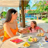 All inclusive com lanches leves Vista superior de uma mesa rústica azul com wraps integrais recheados de folhas em prato amarelo, dois mini bolinhos em travessa branca, um copo de suco verde, uma tigela de pão de queijo e uma salada de frutas; uma mão pega um pão de queijo.