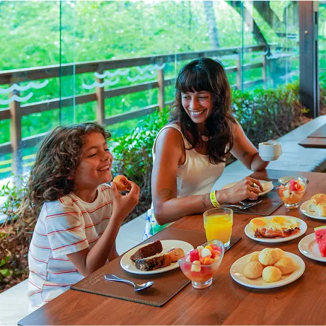 Restaurante galés Três pessoas sorrindo em uma mesa de café da manhã no Restaurante Galés, com grandes janelas de vidro ao fundo revelando área verde. Sobre a mesa há pratos com frutas, melancia, ovos com bacon, pães de queijo, bolos, iogurte com frutas, suco de laranja e xícaras de café; um card com o número 25 aparece ao centro.