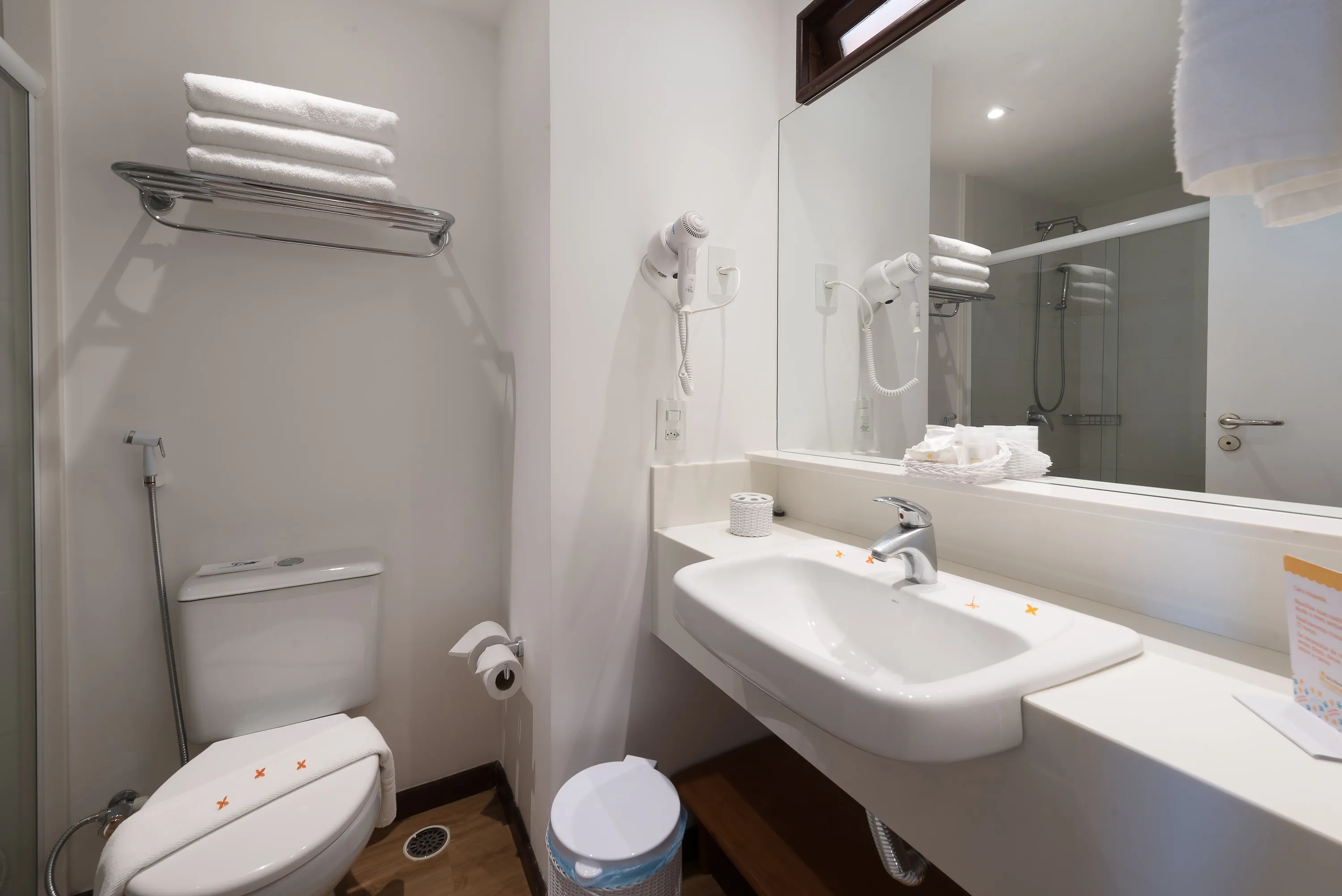 Baño de alojamiento estándar Baño de alojamiento estándar con zona de baño, sanitarios, ducha con control de temperatura, decoración, artículos de aseo O Boticário, toallas, espejo grande y secador de pelo.