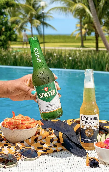 Salinas Maragogi All Inclusive Resort À beira de uma piscina azul, uma mão segura uma garrafa de cerveja Spaten; ao lado, uma Corona long neck, tigelas com camarão e salada sobre uma toalha estampada, óculos de sol e chapéu de palha; ao fundo, palmeiras e o mar desfocados.