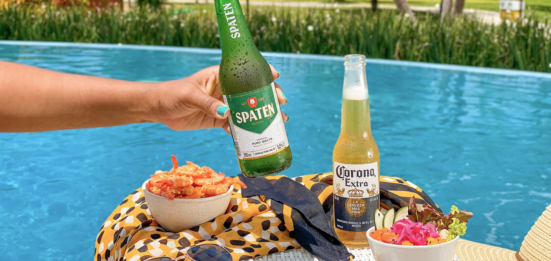 Salinas Maragogi All Inclusive Resort À beira de uma piscina azul, uma mão segura uma garrafa de cerveja Spaten; ao lado, uma Corona long neck, tigelas com camarão e salada sobre uma toalha estampada, óculos de sol e chapéu de palha; ao fundo, palmeiras e o mar desfocados.