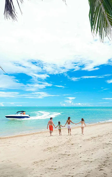 Maragogi Família caminhando pela praia de Maragogi com vista para o mar e um barco ao fundo.