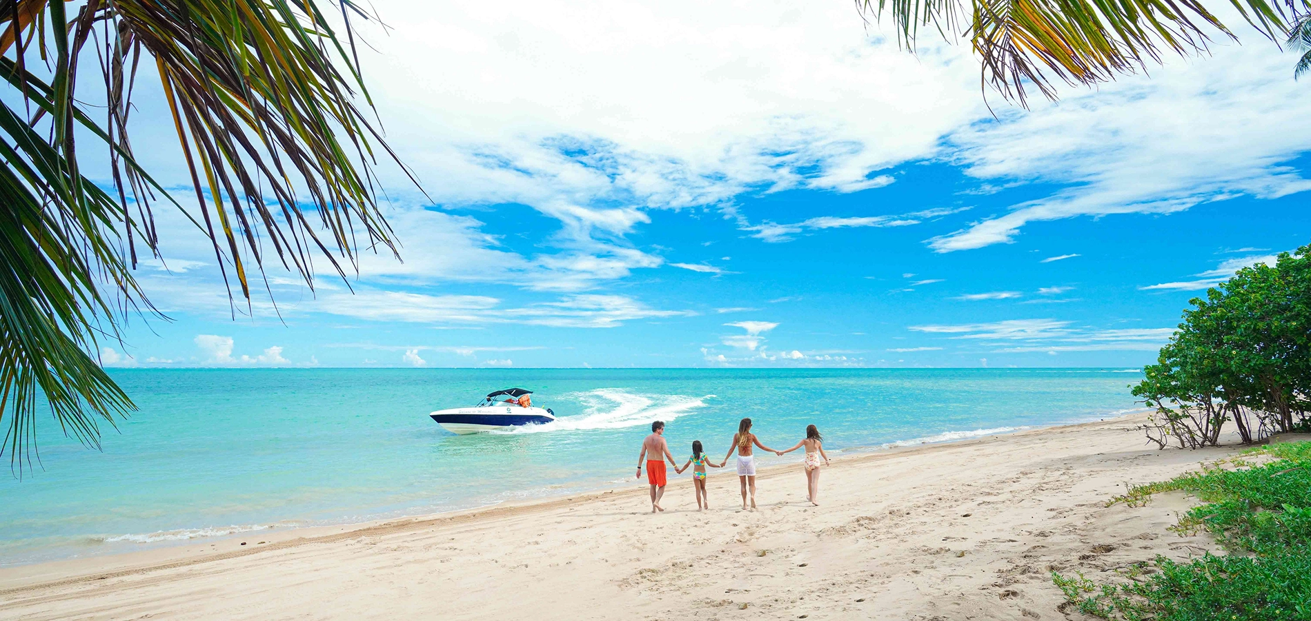 Maragogi Família caminhando pela praia de Maragogi com vista para o mar e um barco ao fundo.