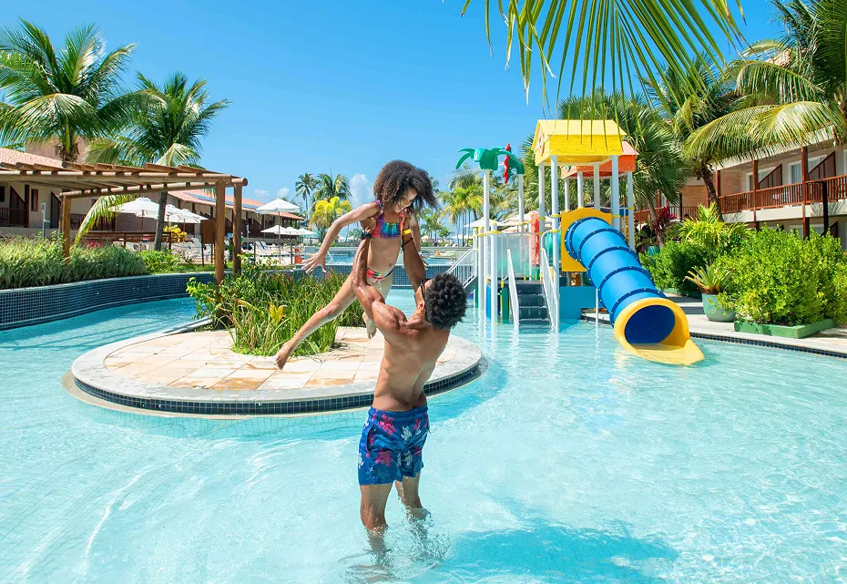Piscina infantil do Salinas Maceió Pai e filha se divertem na piscina infantil do Salinas Maceió, com o pai levantando a criança no ar. Ao fundo, um toboágua colorido e coqueiros completam a cena de férias em família.
