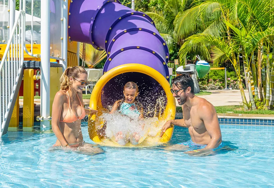 Diversão em família com tobogã, sorrisos e mergulhos Criança saindo de tobogã colorido e sendo recebida por dois adultos sorridentes em piscina de parque aquático, com vegetação tropical ao fundo e céu azul.