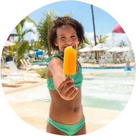 Brincar, refrescar e repetir no melhor resort para família Criança sorridente em traje de banho verde segurando picolé amarelo em frente a um parque aquático com fontes, palmeiras e céu azul.
