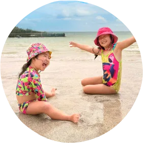 No melhor resort a infância é vivida com liberdade Duas crianças sorridentes sentadas na areia da praia, usando trajes de banho coloridos e chapéus, com mar calmo e céu azul ao fundo.