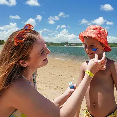 Praia do resort Salinas Maceió Mãe aplicando protetor solar no rosto do filho na praia do Salinas Maceió. Ambos estão sorrindo em um dia ensolarado, com o mar calmo e o céu azul ao fundo.