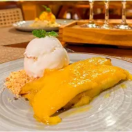 Jantar All Inclusive Prato fundo com arroz, molho cremoso amarelo, legumes grelhados e farofa; mão segura um garfo. Ao fundo, taça de vinho branco e salão de restaurante desfocados.