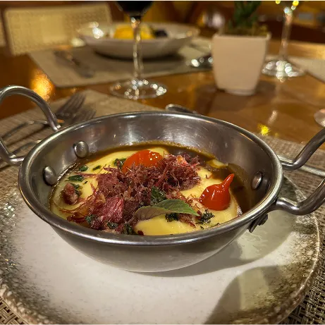 Sobremesa All Inclusive Prato com banana quente coberta por queijo ao lado de uma bola de sorvete e folha de hortelã; ao fundo, três taças de licor sobre bandeja de madeira e salão de restaurante desfocado.