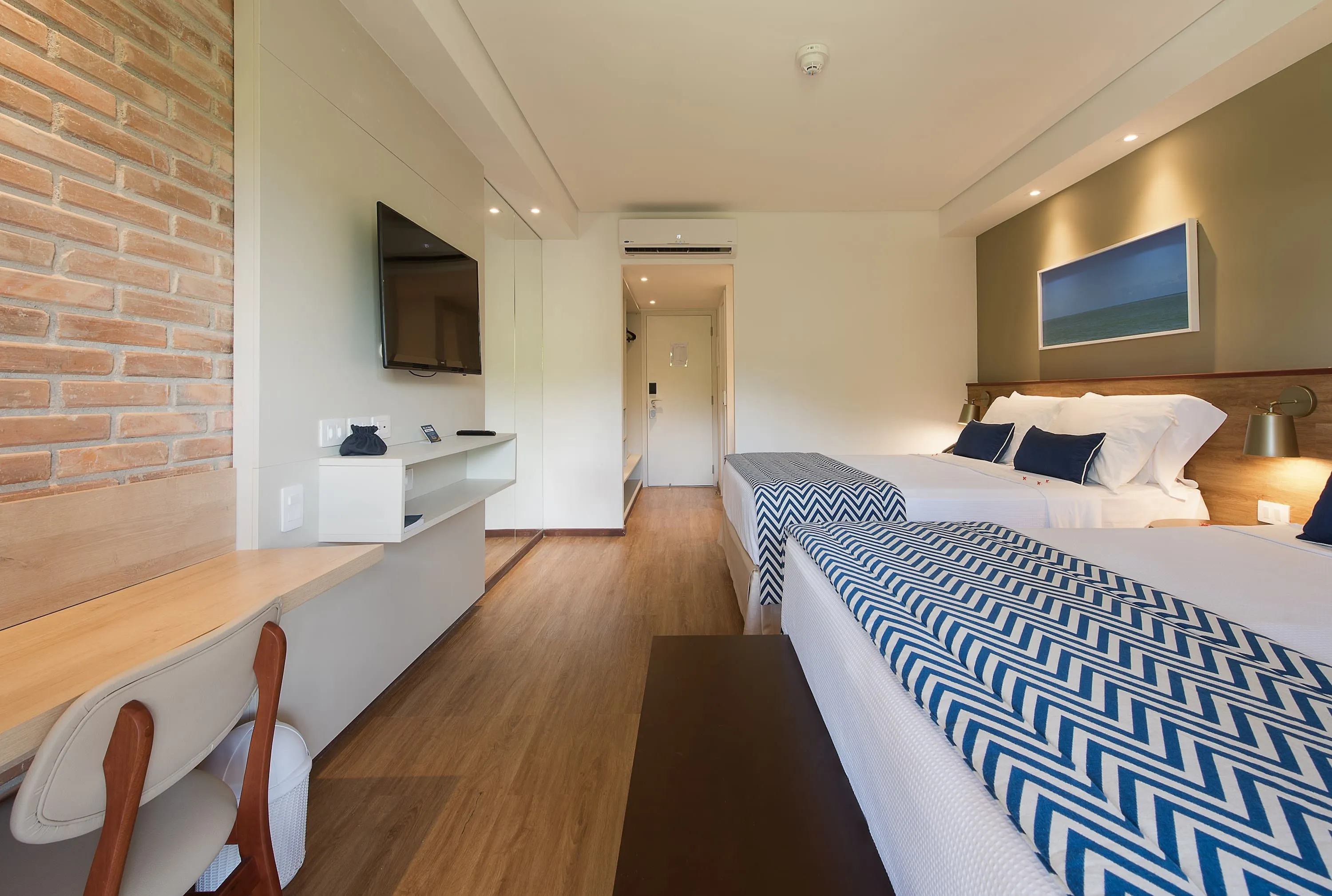 apartamento Playa Jardin Interior del alojamiento Playa Jardín, con dos camas queen, decorado con imágenes de las playas de Maragogi y paredes de ladrillo, además de una mesa con sillas, espejos, armario y puerta de entrada.