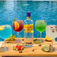 Drinks All Inclusive Cinco coquetéis coloridos à base de gin sobre tábua de madeira ao lado de uma piscina; garrafa de Gordon’s ao centro; copos nas cores azul, vermelho, verde e transparente com guarnições de frutas e especiarias; morangos, kiwi, uvas e anis estrelado na tábua; ao fundo palmeiras, jardim e faixa de praia em dia ensolarado