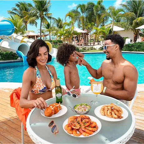 Resort All Inclusive Família à beira da piscina do resort saboreando petiscos e bebidas; ao fundo toboáguas azuis, palmeiras e guarda-sóis em clima tropical