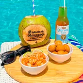 Resort All Inclusive Coco verde com canudo listrado e logo “Salinas Maragogi”, garrafa long neck, tigelas com camarões ao molho e bolinhos, óculos de sol e tábua de madeira sobre espreguiçadeira à beira da piscina azul