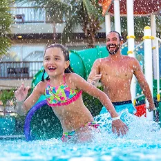 Diversão em família no parque aquático Família se divertindo no parque aquático do Salinas Maceió. Em primeiro plano, uma menina corre sorrindo na água, enquanto seus pais a seguem, rindo, em um momento de pura alegria e descontração.