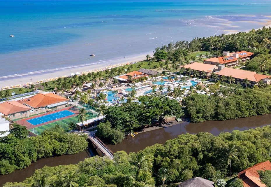 Entre o rio e o mar, o melhor resort na praia Vista aérea do Salinas Maragogi com praia de águas turquesa ao fundo, rio cercado por vegetação tropical, piscinas, quadras e estrutura integrada à natureza.