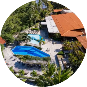 Conforto, natureza e diversão em família entre o mar e o verde. Vista aérea de resort tropical com piscinas, prédios de telhado vermelho, vegetação abundante e canal de água ao lado.