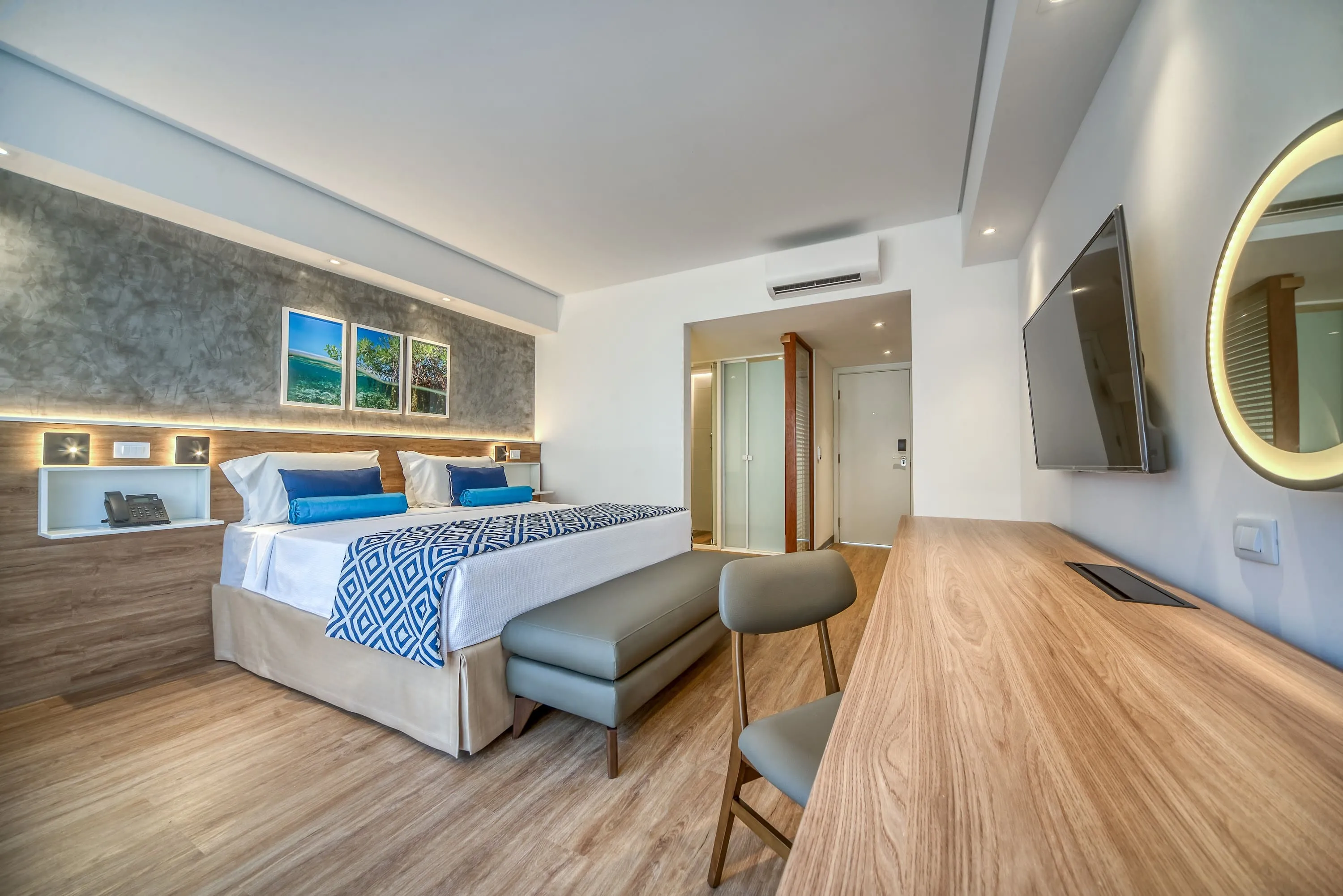 habitación exclusiva den salinas maragogi Configuración del alojamiento en el Estándar Vitória-Régia con cama queen size, mesa y silla con espejo redondo iluminado, armoniosa decoración de madera, lámparas y marcos con fotografías de las playas de Maragogi. También puede ver el baño y la entrada del apartamento.