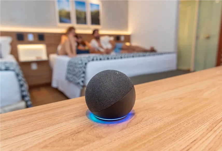 acomodação tecnológica salinas maragogi Alexa integrada ao Standard Vitória-Régia, sobre mesa de madeira, em frente às camas onde uma família repousa.