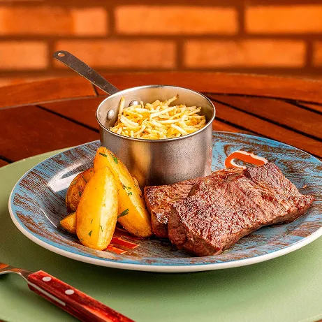 Lanche all inclusive Prato com hambúrguer artesanal com queijo, alface e tomate, batatas fritas e potinho de molho, sobre mesa de madeira; ao fundo, parede de tijolos.