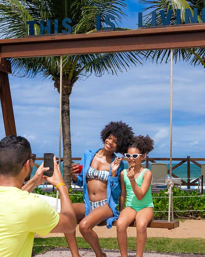 resort maceió Pai bate foto de esposa e filha no balanço do deck do Salinas Maceió, com o mar e céu azul da praia de Ipioca ao fundo.