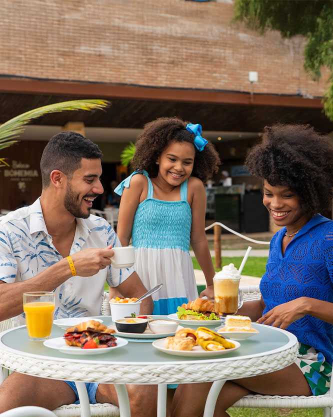 férias em família maceió Família feliz desfrutando um lanche da tarde em frente ao Café Canoas e ao Boteco Bohemia no Salinas Maceió, sentados ao lado da mesa, onde nela tem pratos diversos (frutas, pãeszinhos, sobremesas) , suco e café gelado.