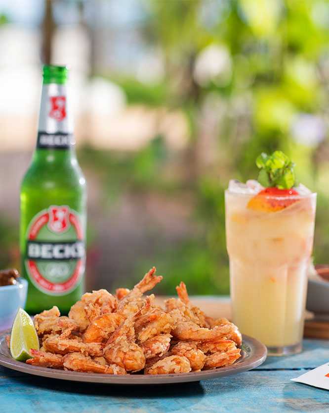 resort all inclusive Diversos petiscos (camarão empanado, carnes e salada) sobre mesa azul, ao lado de drinks e cerveja Becks, em frente à vegetação nativa da praia de Ipioca, onde está o Salinas Maceió.