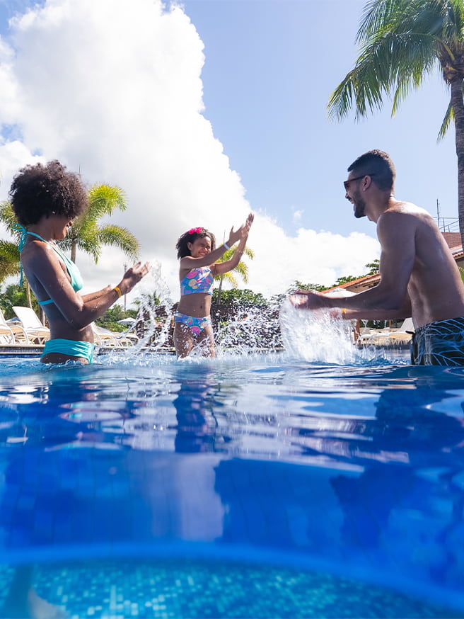 Diversão em família nas piscinas do Salinas Maceió Família se divertindo na piscina do Salinas Maceió com pai, mãe e filha jogando água pra cima. Ao redor toda a estrutura do resort, com espreguiçadeiras, coqueiros e guarda-sóis.