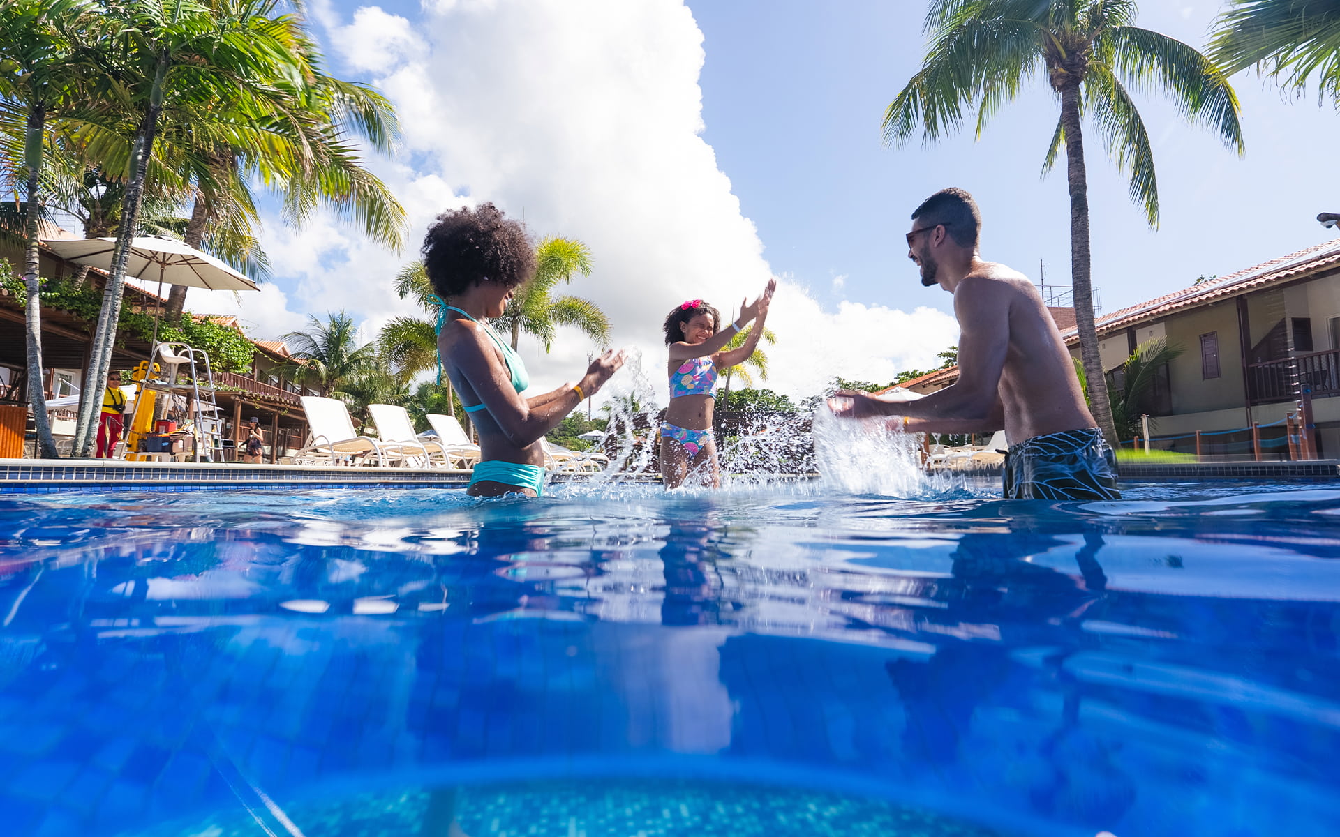 Diversão em família nas piscinas do Salinas Maceió Família se divertindo na piscina do Salinas Maceió com pai, mãe e filha jogando água pra cima. Ao redor toda a estrutura do resort, com espreguiçadeiras, coqueiros e guarda-sóis.