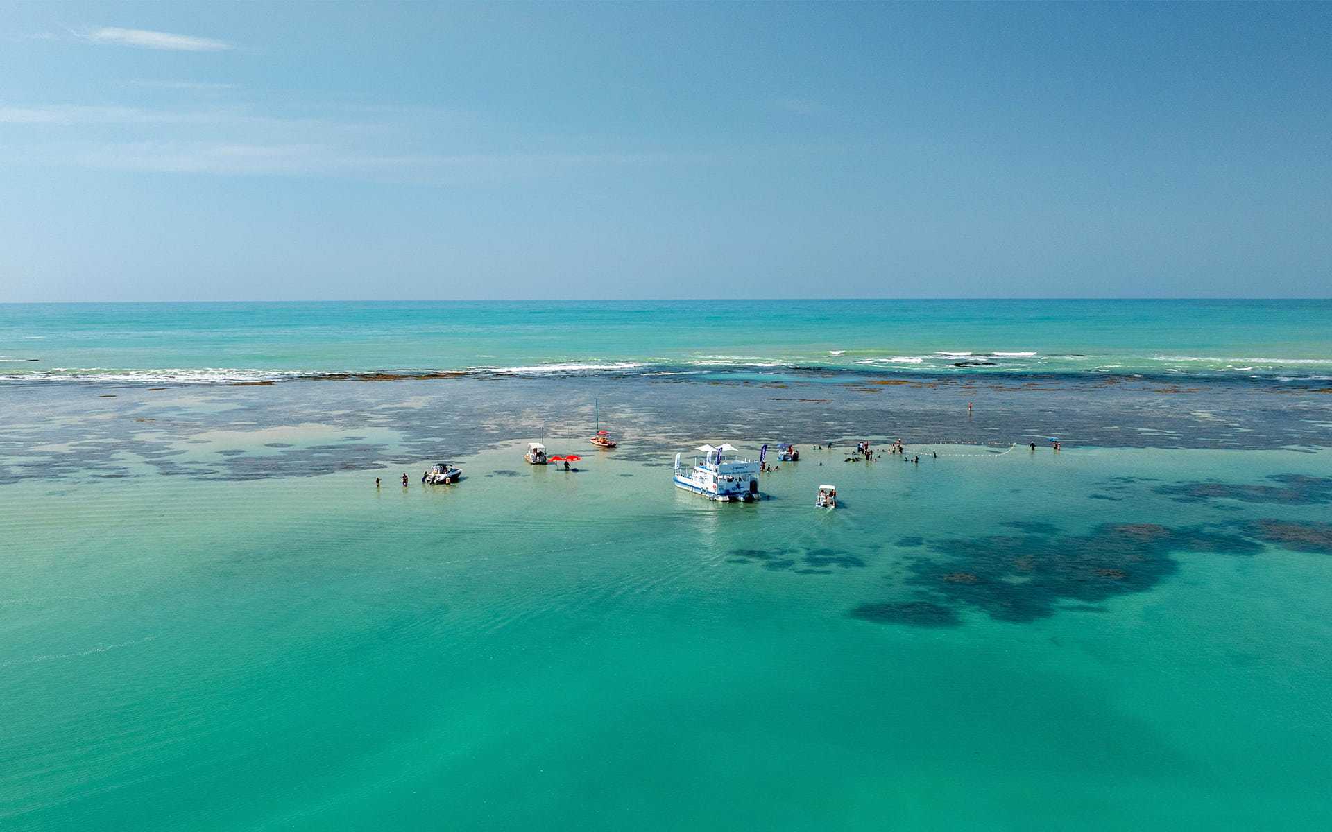 Praia de Pajuçara com águas cristalinas Vista aérea da Praia de Pajuçara com mar cristalino, recifes de corais visíveis e algumas embarcações atracadas nas piscinas naturais.