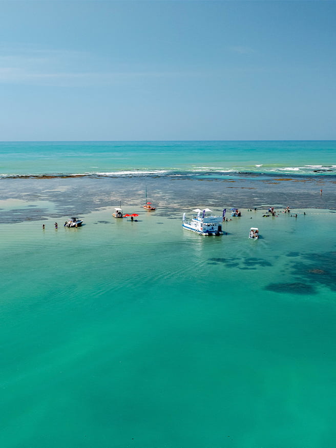 Praia de Pajuçara com águas cristalinas Vista aérea da Praia de Pajuçara com mar cristalino, recifes de corais visíveis e algumas embarcações atracadas nas piscinas naturais.